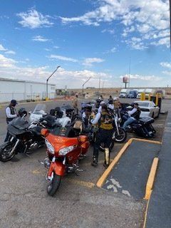 Motorcycles and riders in parking lot. Orange, black, and white bikes with people wearing vests, under blue sky.