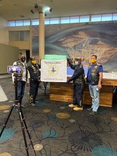 Four people holding a large, decorated sign. Filming setup in front of a mural.