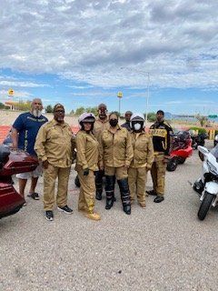Group of people in tan outfits posing by motorcycles in a parking lot under a cloudy sky.
