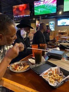 Group of people at a bar, eating food and watching TVs. One man wears a hat and mask.