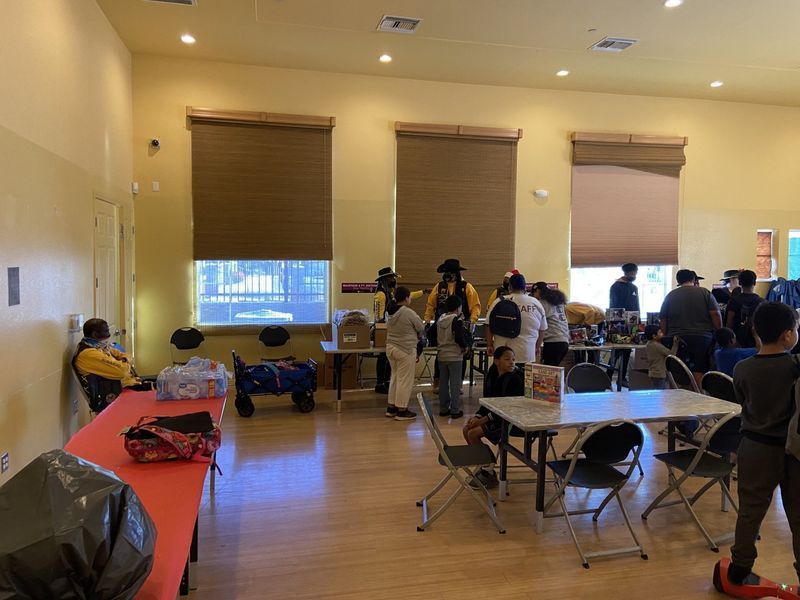 People at tables in a community room; some are browsing items for sale. Three windows with shades. Wooden floor.