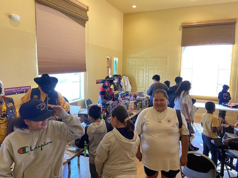 People browsing tables with items, indoors. Several children, window with shade. Some wearing hats.