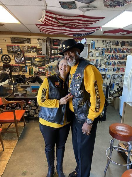 Two people in black vests and yellow shirts, posing in a shop with motorcycle parts, smiling.