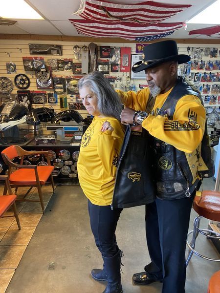 Man zips a black leather vest on a woman wearing a yellow shirt. They are in a shop with flags and motorcycle parts.