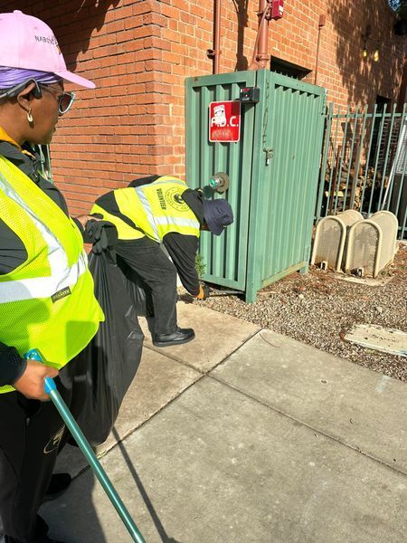 Two people in reflective vests near a green metal container, one bending down with a black bag.