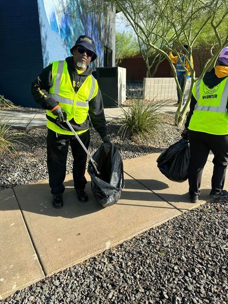 Two people in reflective vests picking up trash outdoors. One holds a grabber, the other a trash bag.