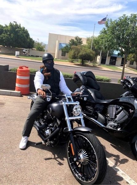 Person on a black motorcycle, near a parked, black motorcycle. Outdoors, sunny day.