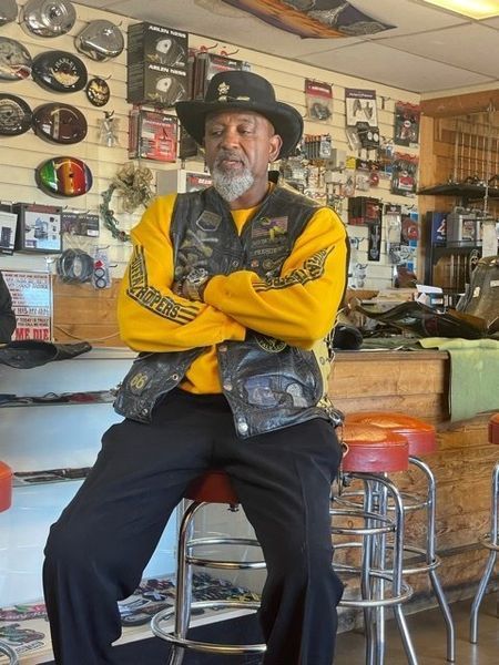Man in yellow shirt and vest sits in a shop. He has arms crossed and a serious expression.
