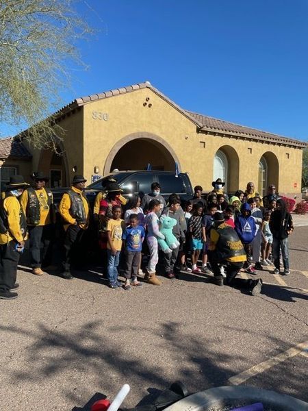 Group of children and adults pose in front of a building. Some adults wear yellow and black. Sunlight.