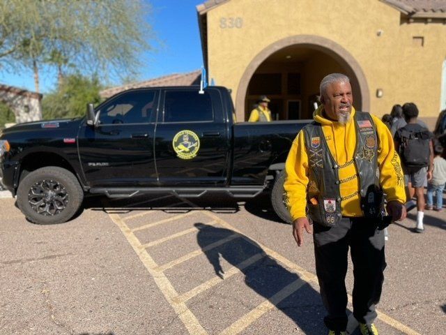 Man in yellow vest walks near black truck with logo, building in background.