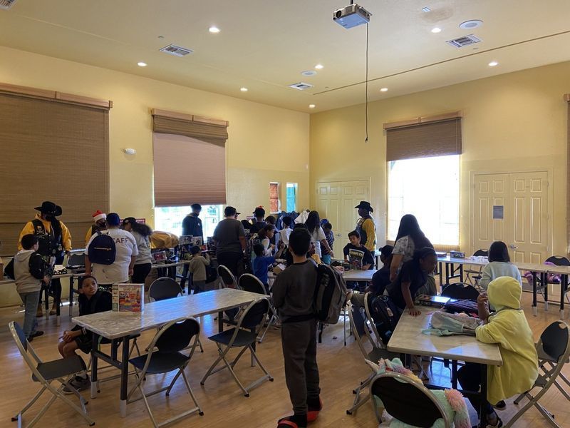 People in a brightly lit room with folding tables, participating in an event. Some are seated, some are standing.