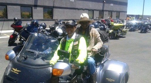 Two people on a trike motorcycle, others in background, parked outdoors. Both wear jackets.