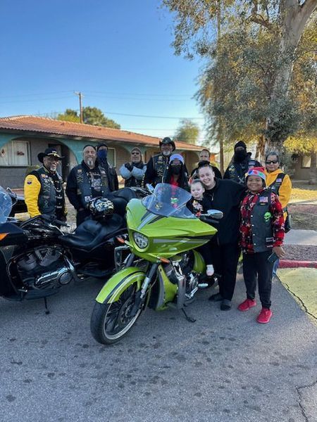 A group of people poses with motorcycles. A bright green bike is in the center. People wear leather vests and jackets. Sunny outdoor setting.