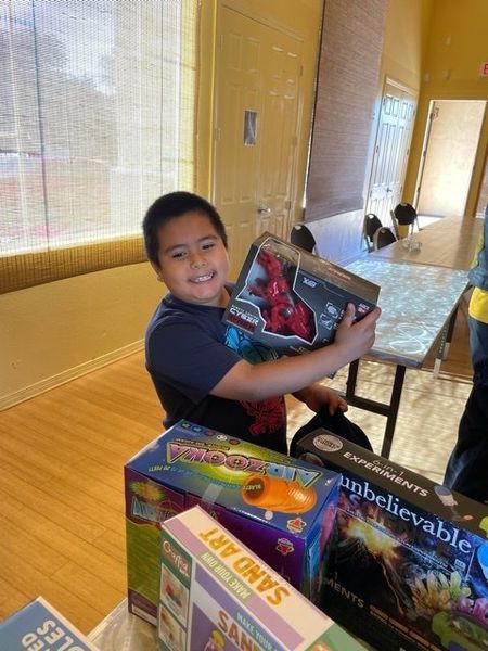 Boy holding a toy, smiling. Surrounded by other toys on a table in a room with a window.