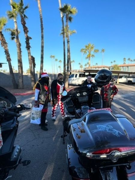 People in festive attire pose next to motorcycles in a sunny parking lot. Palm trees in background.
