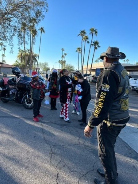 Group gathers near motorcycles, palm trees in the background. Man in hat and vest faces the group.
