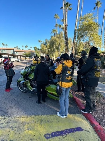 A group of people wearing jackets with patches gather around a green motorcycle outdoors.