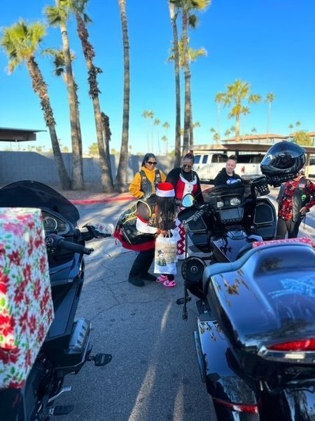 Motorcyclists by bikes; person in Santa hat and pajamas, presents; palm trees and sunny background.
