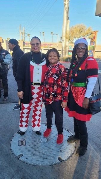 Three people in festive outfits pose outdoors on a sunny day. One wears a Christmas sweater and pants, another a red and black patterned jacket, and a Santa hat.