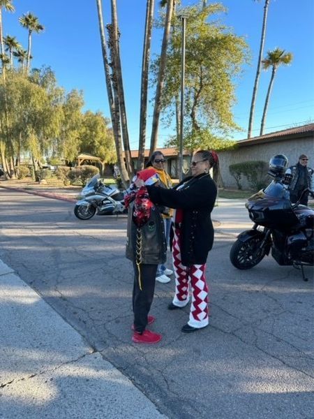 People on a street with motorcycles. One person adjusting another's red and white patterned pants and a hat.