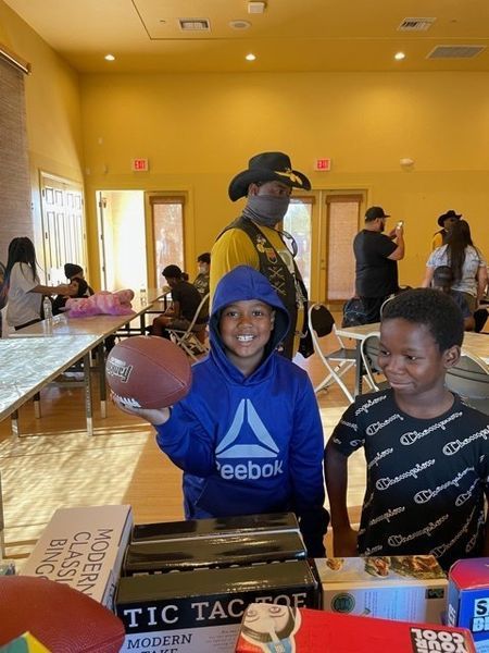 Two boys with a football at a community event; one smiles holding the ball. A man in a cowboy hat is in the background.