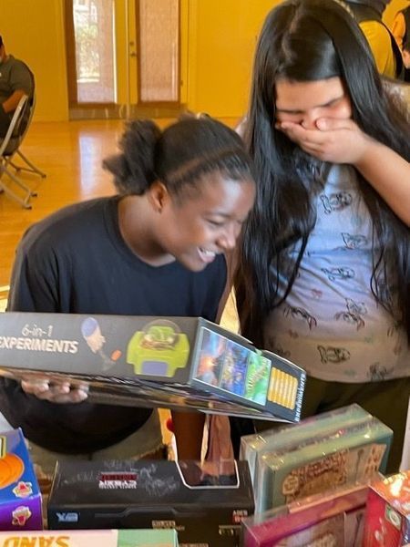 Two people smiling, looking at a box of science experiments. The other person is covering their mouth, with a light blue shirt.