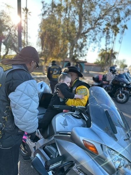 Child held by motorcyclist, hugging on motorcycle. Others gather, sunny day.
