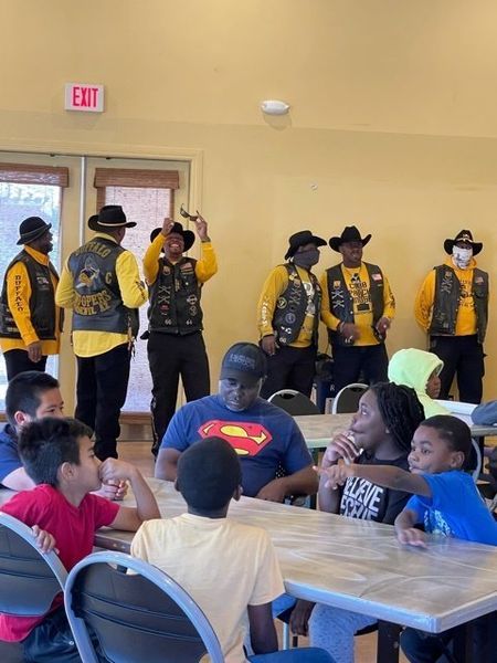 Group of people, some wearing vests and hats, standing near children seated at tables indoors.