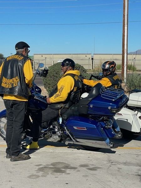 Three people, two on a blue motorcycle, wearing yellow vests with a club logo, standing in a parking lot.