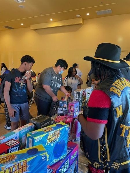 People browsing a table of toys. One person in a vest. Indoors, well-lit room.