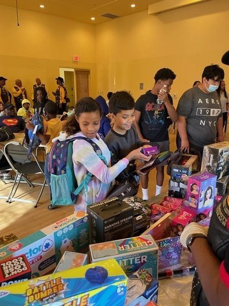 Children selecting toys from a table in a brightly lit room. Other people are in the background.