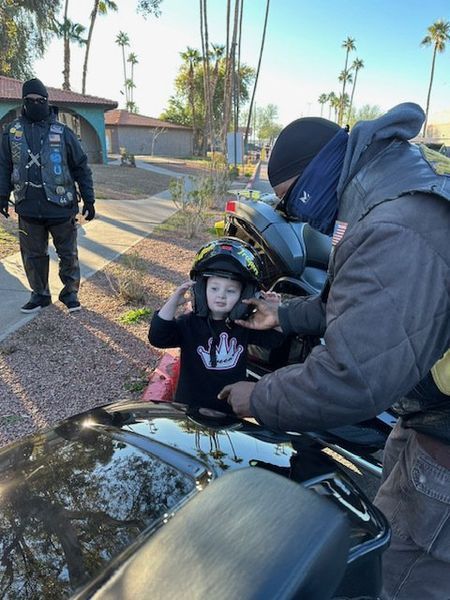 Boy in helmet being helped by a person near a motorcycle. Another person in background.