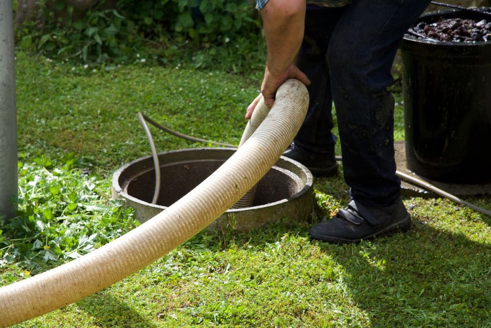 Person in dark pants and shoes holding a large hose over an open septic tank in a grassy yard.