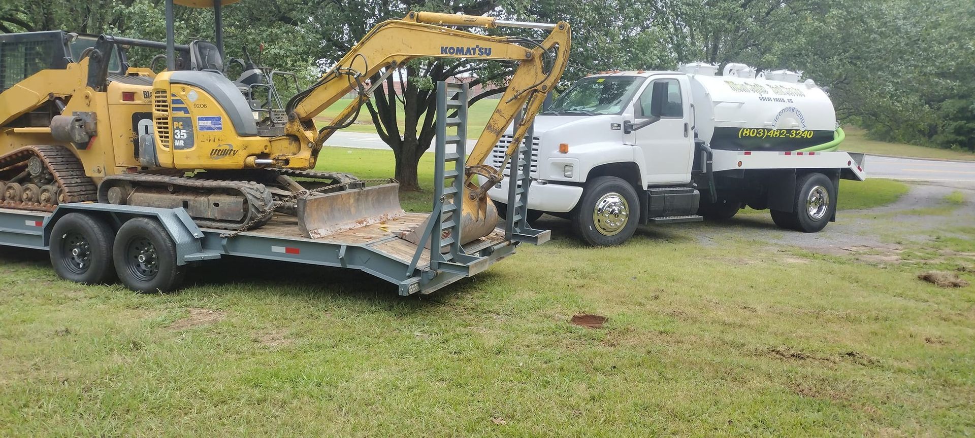 A yellow excavator on a trailer next to a white water tank truck on grass.
