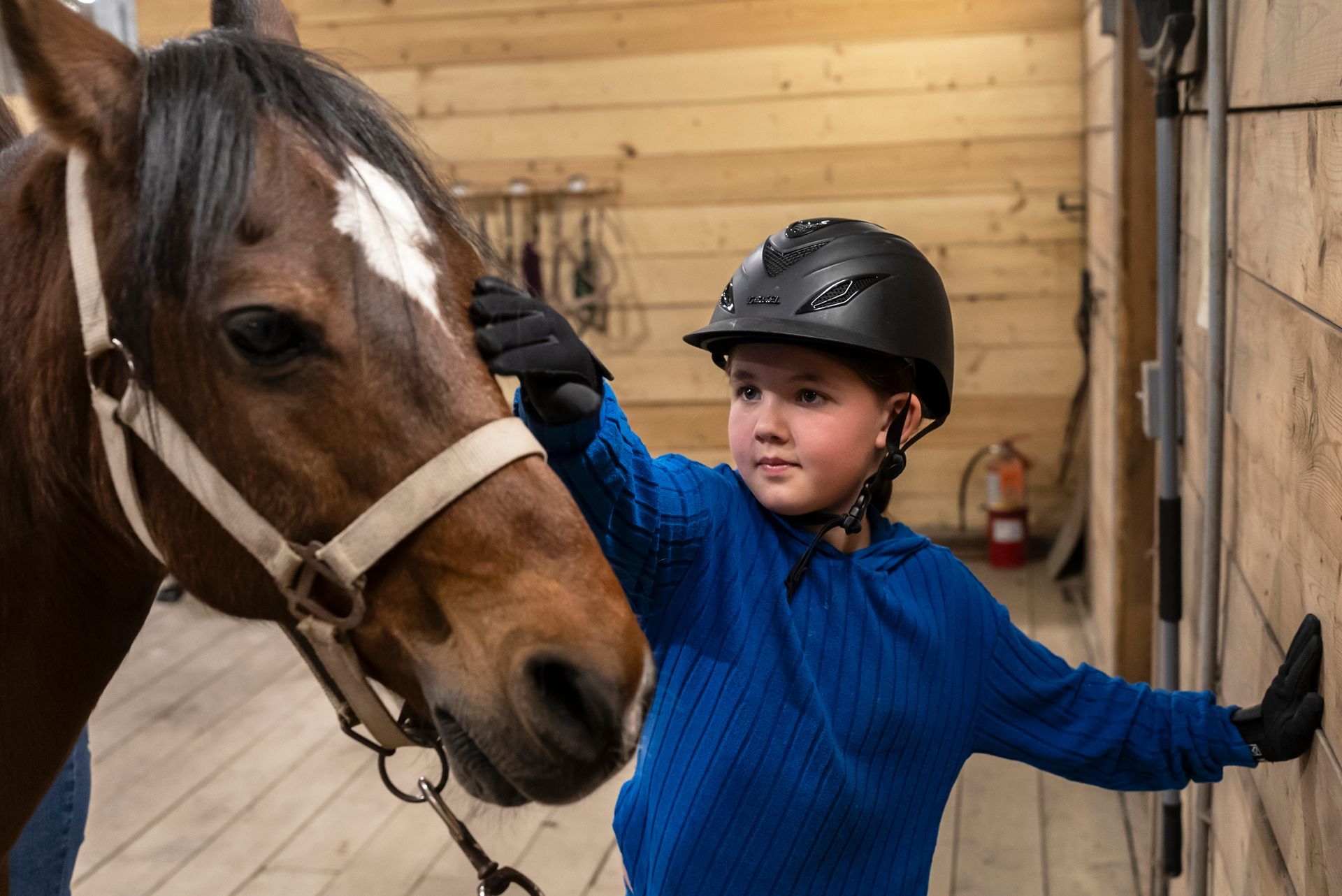 A young girl wearing a helmet is petting a horse in a stable.