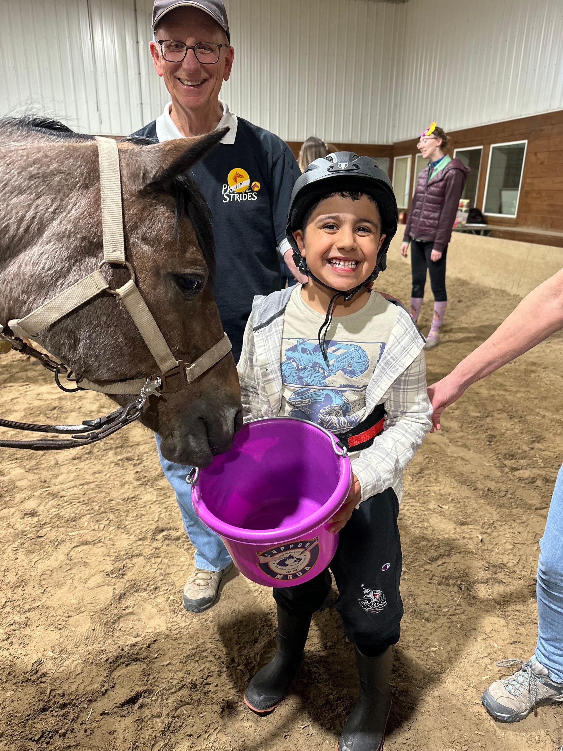 Boy smiles, feeding a horse from a purple bucket. Man smiles beside them in an indoor arena.