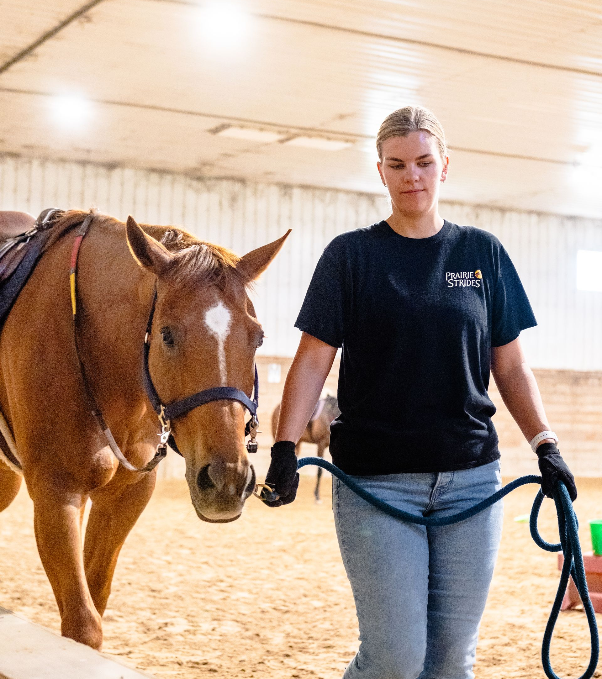 Woman leading a saddled brown horse with a blue lead rope in a barn.