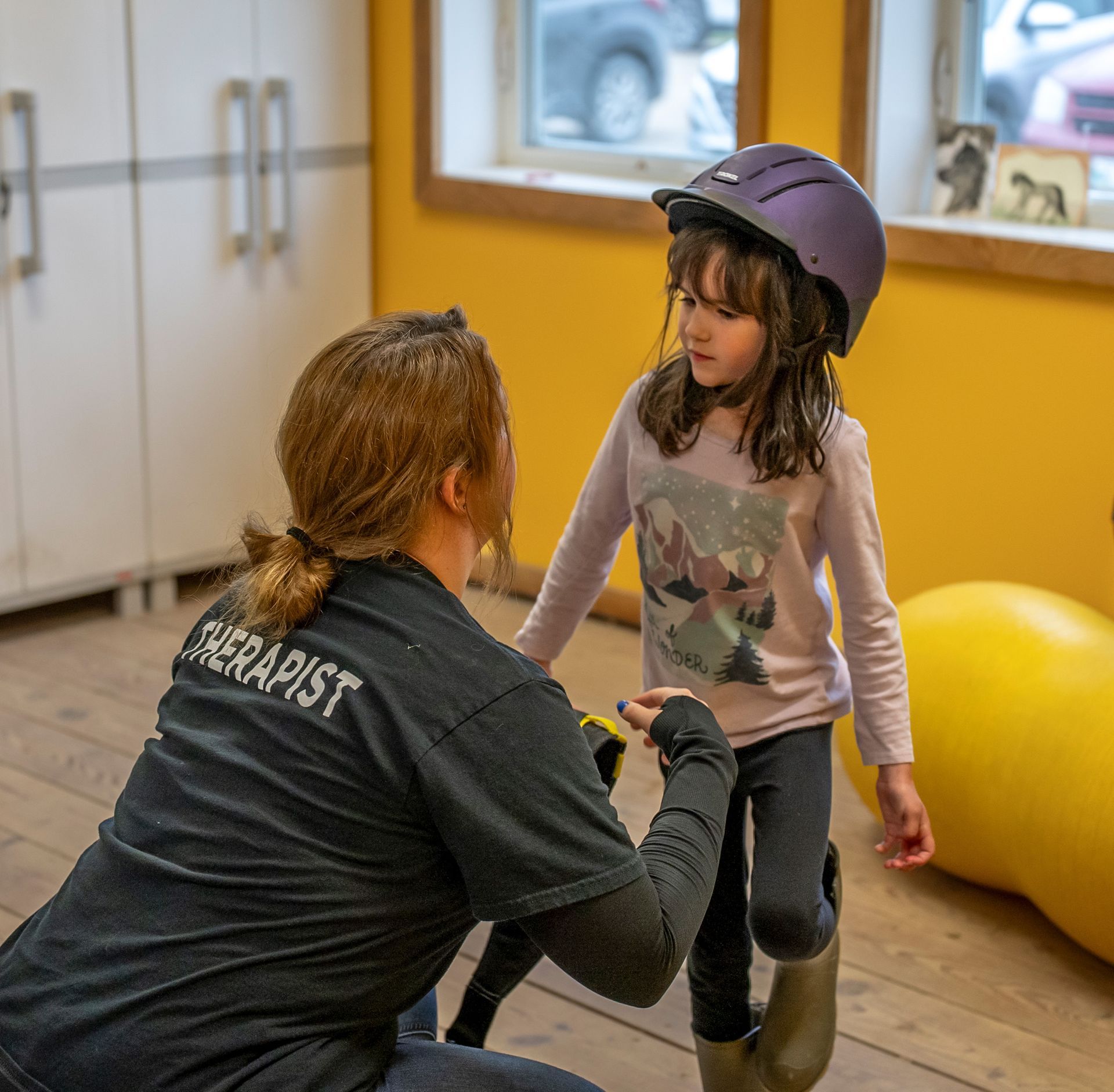 Woman, labeled "Therapist," assists girl in helmet, indoors, yellow wall, white cabinets, window.