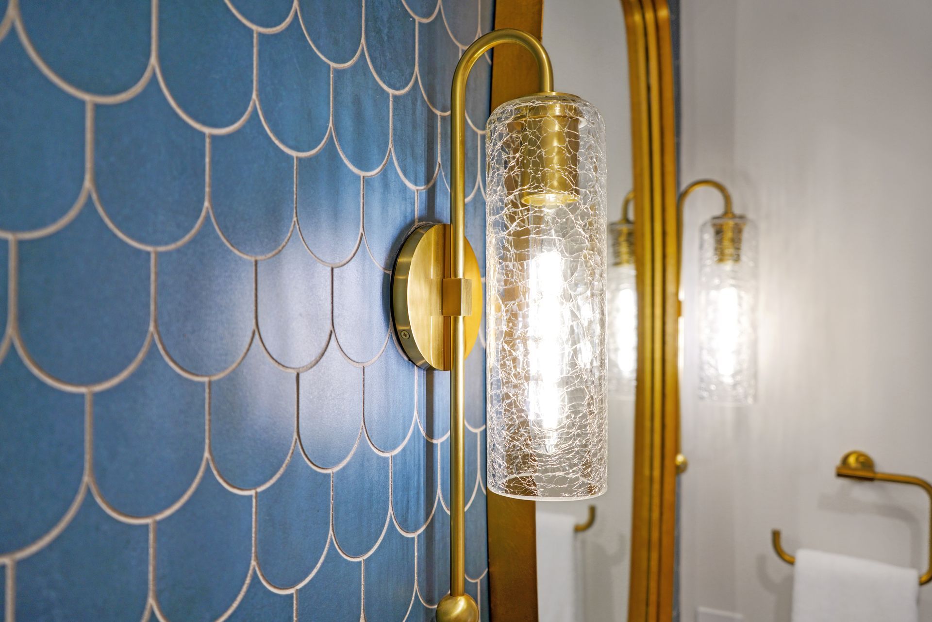 Gold-toned wall sconce with a textured glass shade and lightbulb against a blue scalloped tile wall.