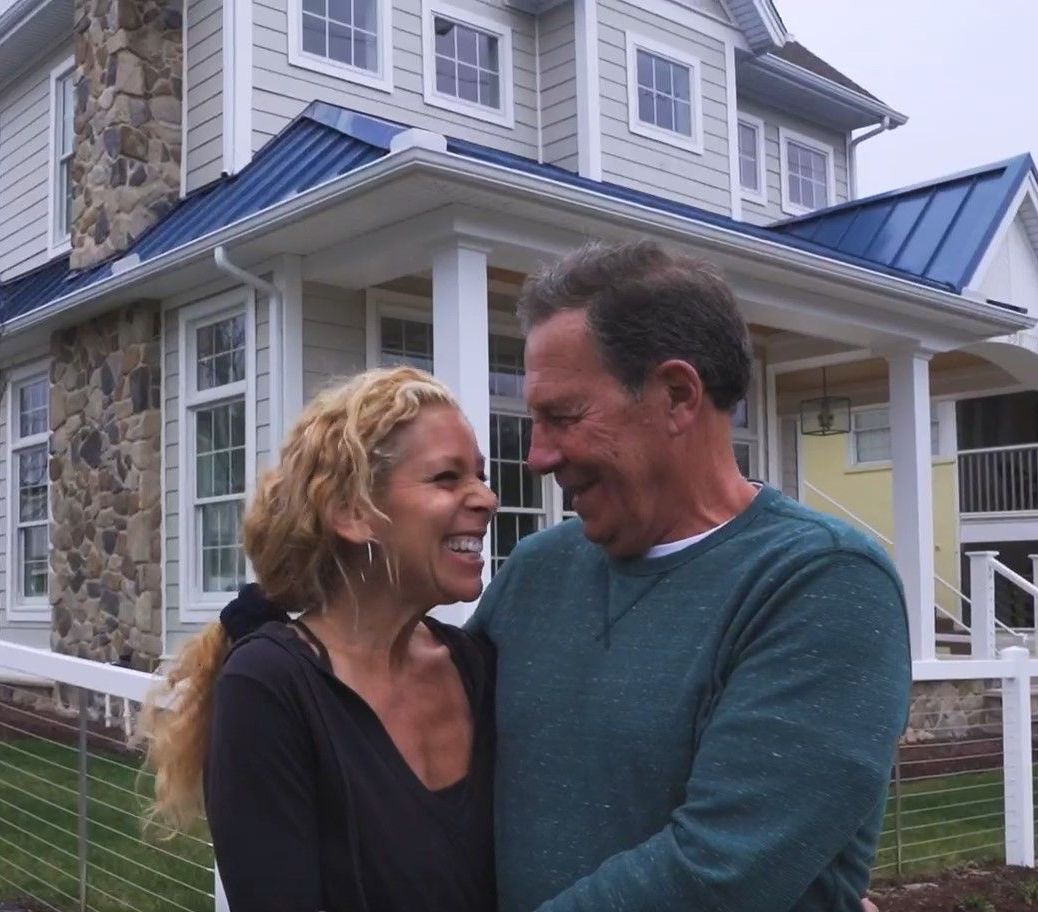 Couple embracing, laughing in front of a large house with blue roof and stone chimney.