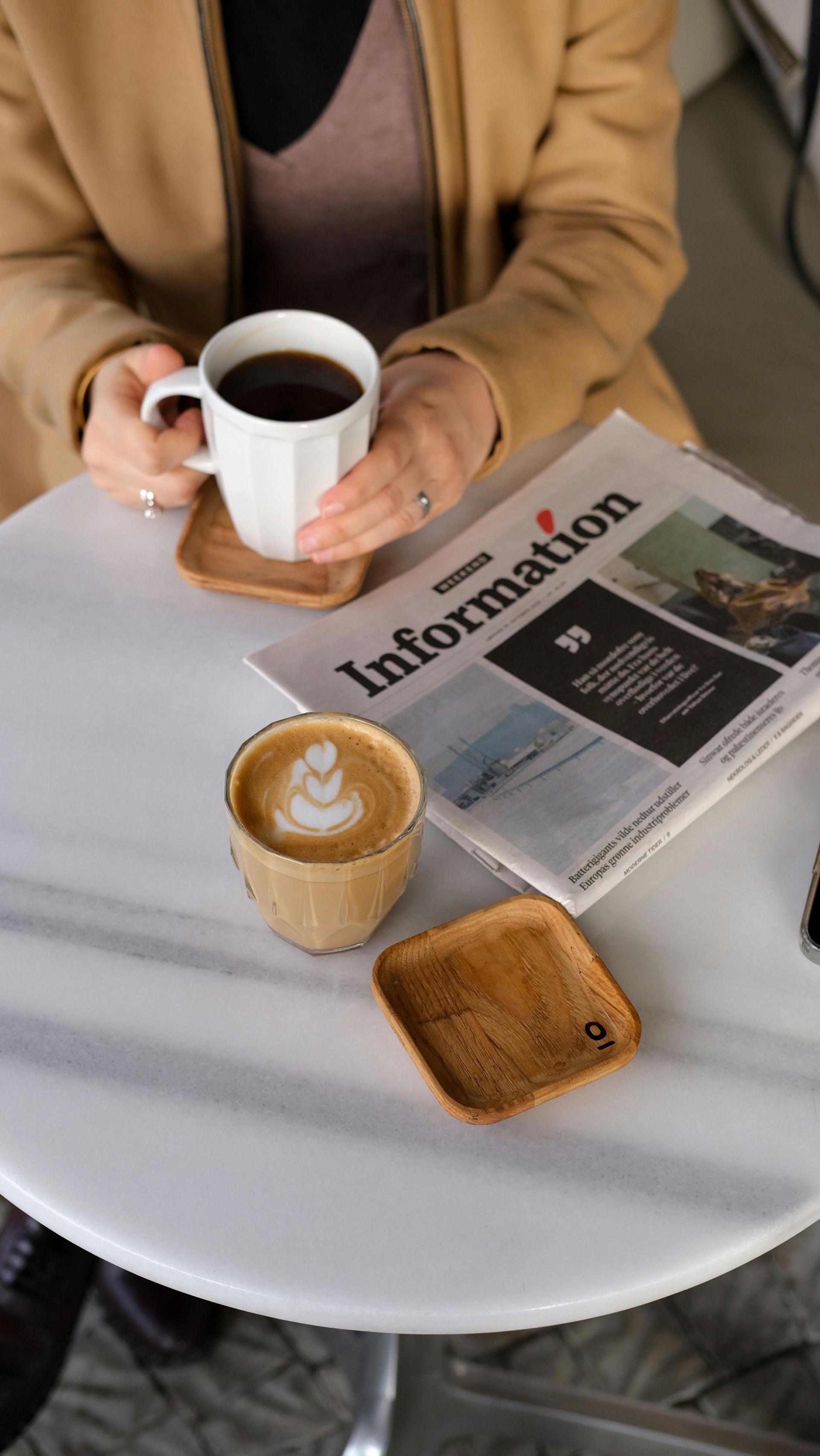 Woman with coffee and newspaper at a table. Cappuccino, black coffee, and wooden coasters.
