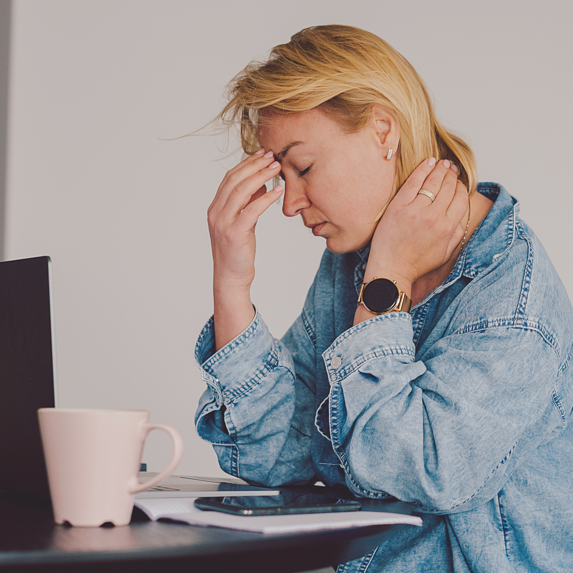 A woman in a denim shirt is sitting at a desk with her head in her hands.