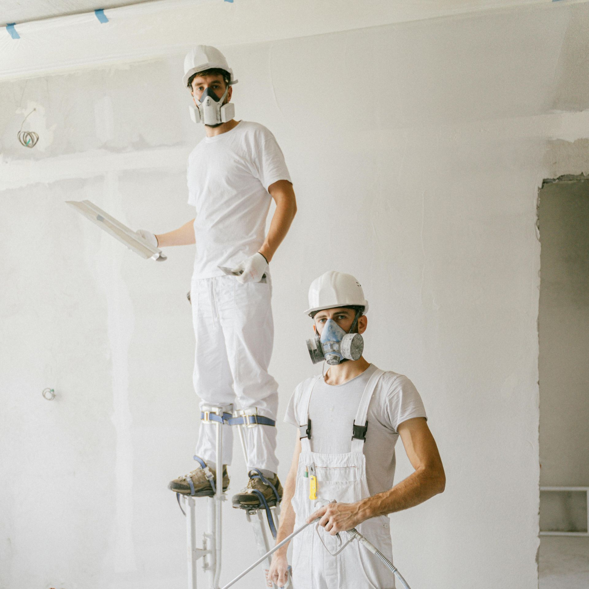 Two drywall contractors wearing masks and stilts in a white room.