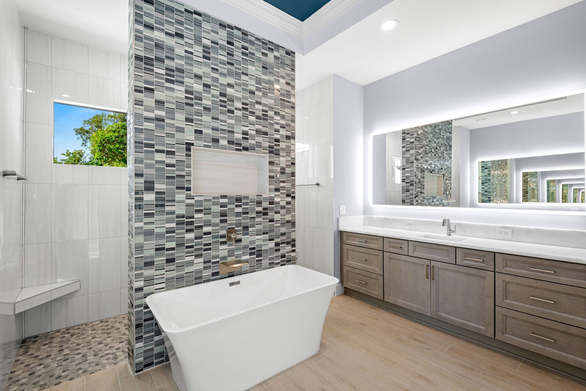 A modern bathroom with a freestanding white tub, tiled accent wall, light wood flooring, and a large illuminated mirror.