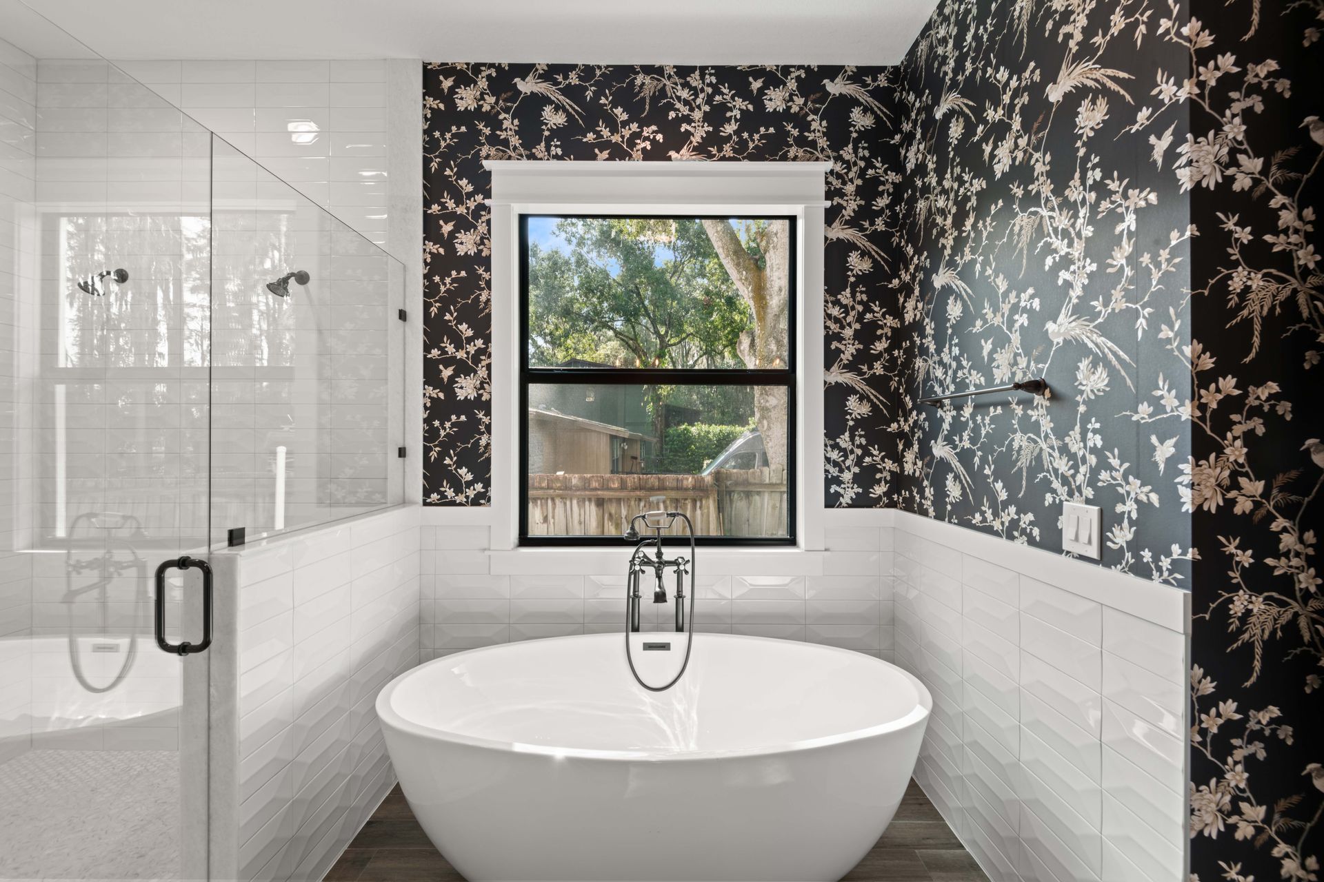 A white freestanding tub sits before a window framed by black floral wallpaper, with a glass shower stall to the left.
