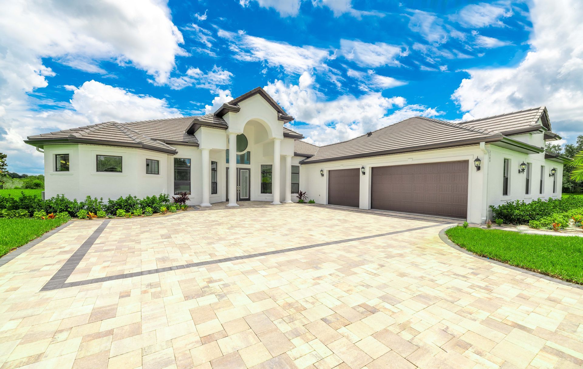Large, white luxury home with beige driveway and garage doors; blue sky with clouds overhead.