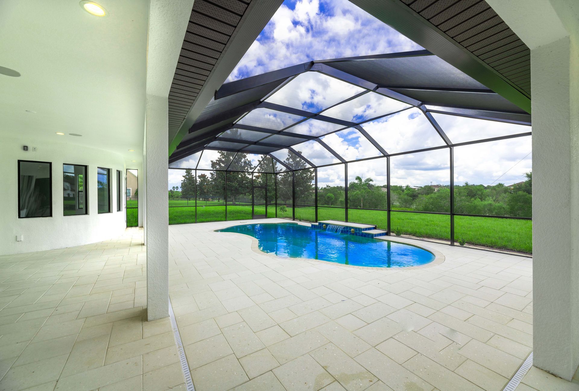 Open-air pool area with blue water, white tile flooring, and a screened enclosure against a bright sky.