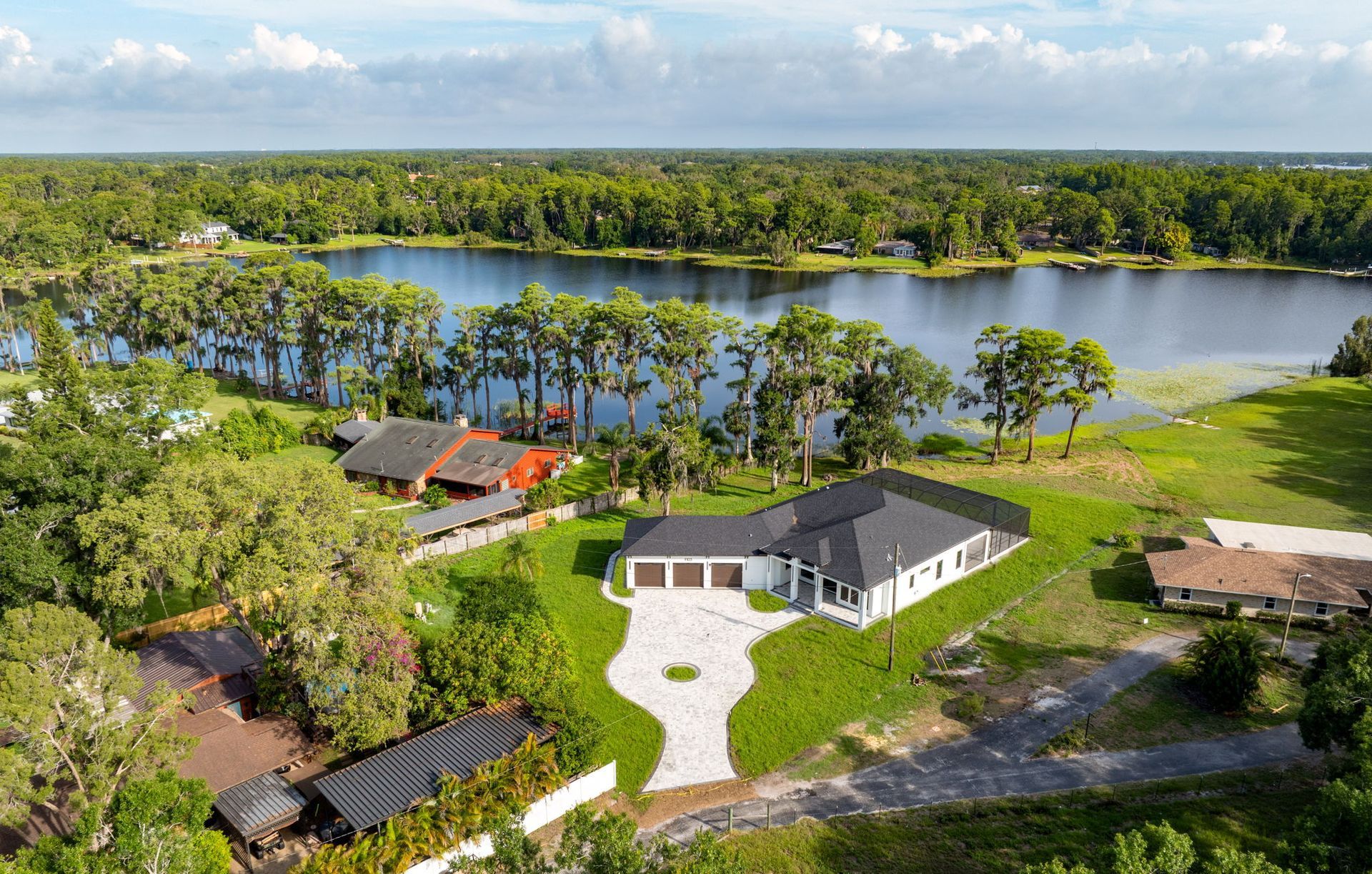 Aerial view of a modern, white house with a black roof next to a lake, surrounded by trees.