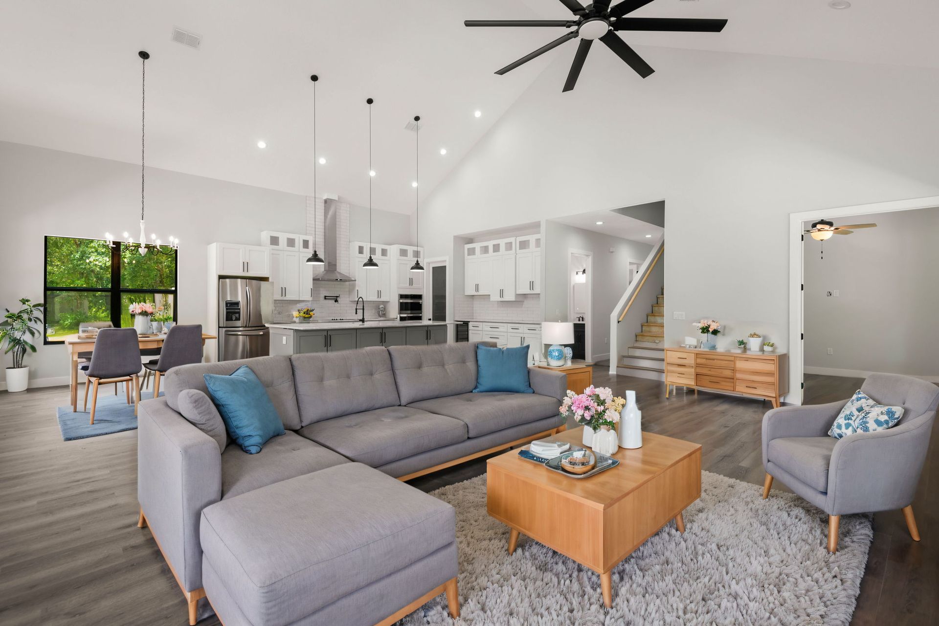 Modern open-concept living room with gray sectional, light wood accents, and kitchen in the background.