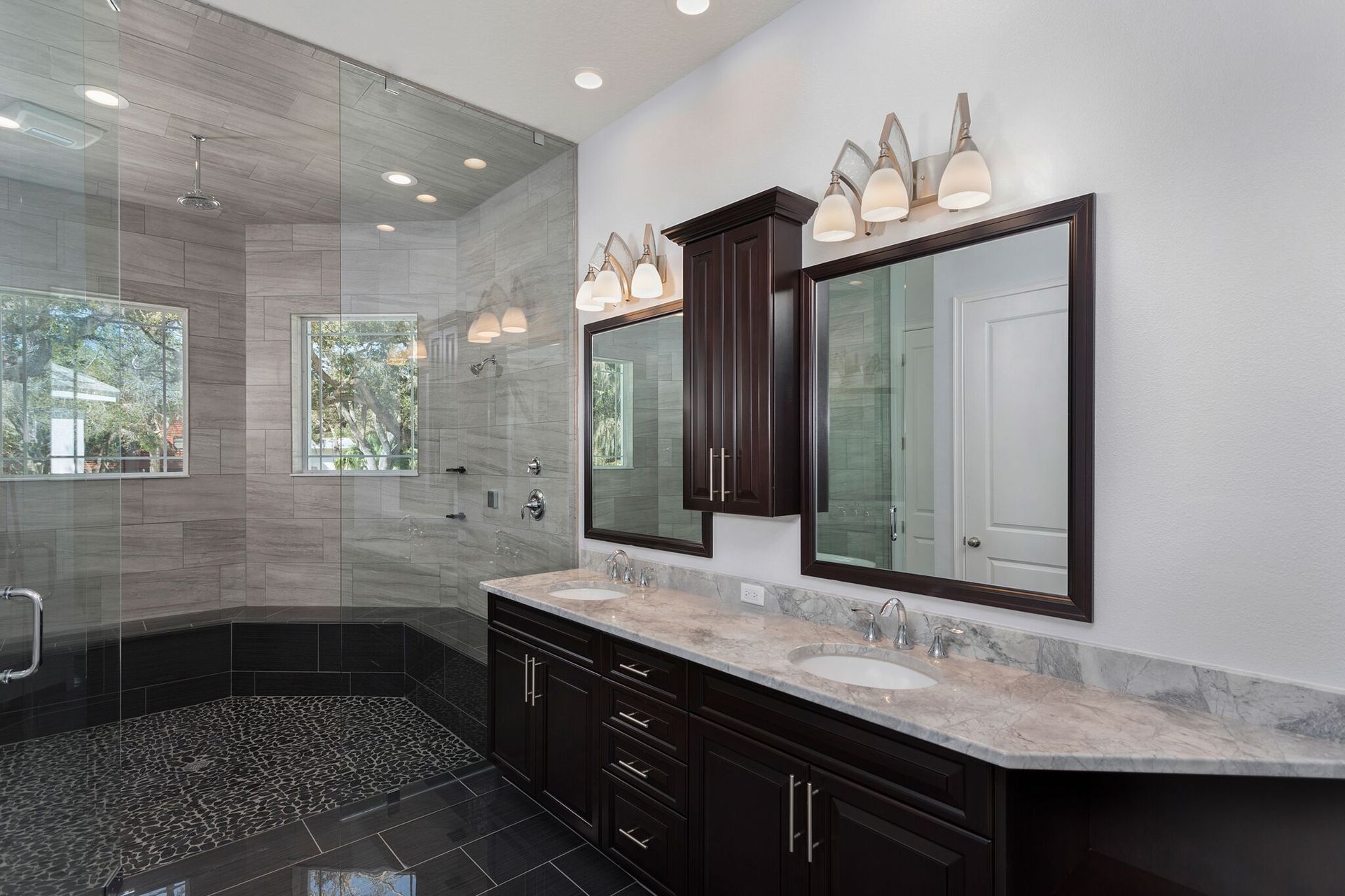 A modern bathroom with a glass-enclosed shower featuring stone floors, dual sinks, dark cabinetry, and matching mirrors.